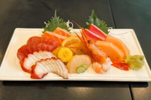 An artfully arranged platter of fresh, raw seafood on a rectangular white ceramic plate, featuring slices of tuna, salmon, octopus, swordfish, scallops, and a whole sweet prawn, garnished with green shiso leaves, lemon slices, a yellow edible flower, and a portion of wasabi.