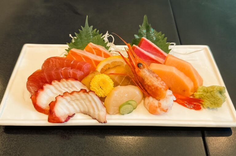 An artfully arranged platter of fresh, raw seafood on a rectangular white ceramic plate, featuring slices of tuna, salmon, octopus, swordfish, scallops, and a whole sweet prawn, garnished with green shiso leaves, lemon slices, a yellow edible flower, and a portion of wasabi.