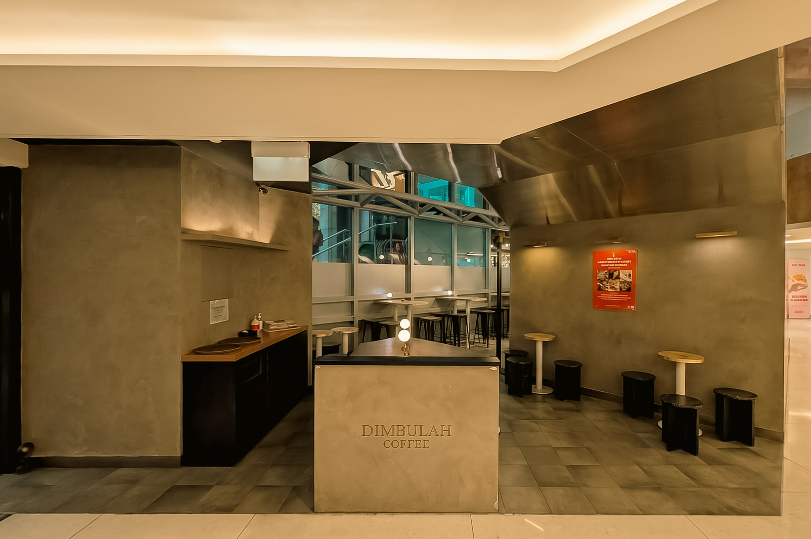 Modern cafe interior featuring smooth, warm-toned gray concrete walls, tile flooring, and metallic ceiling paneling. In the center stands a concrete counter engraved with "DIMBULAH COFFEE." Small round seating tables paired with low black and white stools are positioned along the wall and windowed partition in the background.