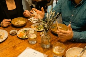 Two people are seated at a wooden table during a meal, surrounded by bowls of food, side dishes, a menu, and glasses of water with lemon slices. A small glass vase containing leafy twigs sits in the center of the table, while the person on the right gestures with their hands while speaking.