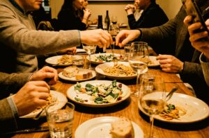 A group of friends gathered around a wooden dining table sharing a meal of pasta dishes, fresh salads, bread, and wine, with one person holding a smartphone above the table.