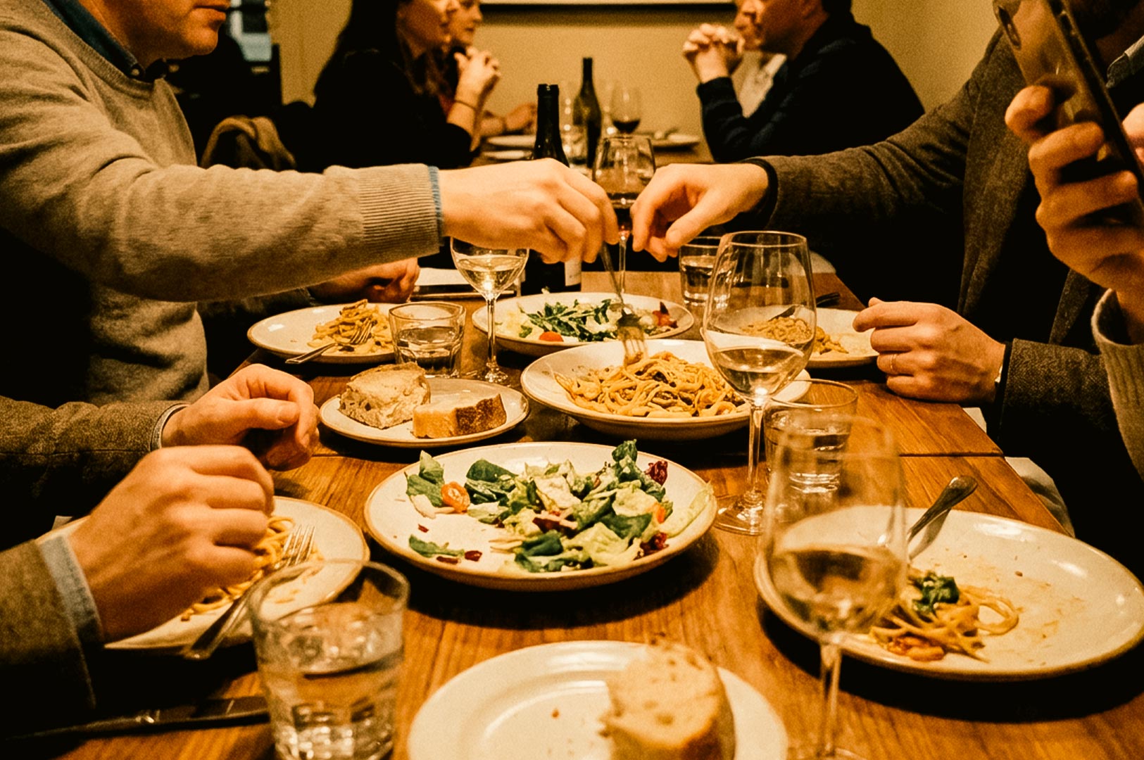 A group of friends gathered around a wooden dining table sharing a meal of pasta dishes, fresh salads, bread, and wine, with one person holding a smartphone above the table.