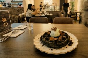 The image features a dark charcoal or chocolate waffle served on a white, scalloped plate on a wooden table in the foreground. The waffle is topped with a scoop of light-colored ice cream and cereal pieces, accompanied by a glass of water, cutlery wrapped in napkins, and a menu stand. In the background, the softly lit interior of a cafe or restaurant shows other tables, patrons seated and talking, and staff members working near a counter.