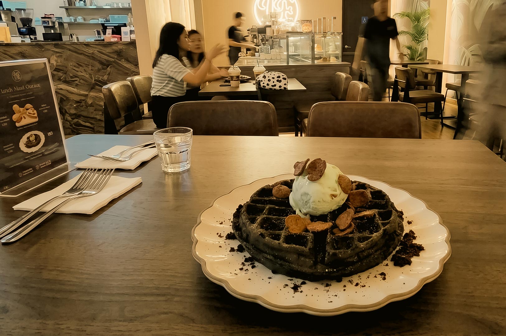 The image features a dark charcoal or chocolate waffle served on a white, scalloped plate on a wooden table in the foreground. The waffle is topped with a scoop of light-colored ice cream and cereal pieces, accompanied by a glass of water, cutlery wrapped in napkins, and a menu stand. In the background, the softly lit interior of a cafe or restaurant shows other tables, patrons seated and talking, and staff members working near a counter.
