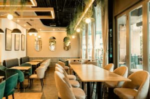 A bright, modern restaurant interior with a warm and elegant atmosphere. Light wood tables are paired with cream-colored upholstered chairs and deep green velvet seating, while large floor-to-ceiling windows fill the space with natural light. Contemporary pendant lights and geometric ceiling fixtures hang alongside cascading greenery, creating a fresh, inviting ambiance. Framed artwork and oval mirrors decorate the walls, and the neatly arranged tables provide a spacious, comfortable setting suitable for casual dining, business lunches, or group gatherings.
