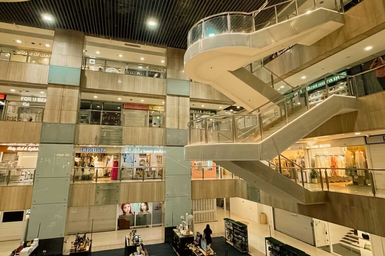 A low-angle view looking up at a multi-level indoor shopping mall atrium featuring sweeping helical staircases with glass railings, surrounded by tiers of boutique store fronts and kiosks.