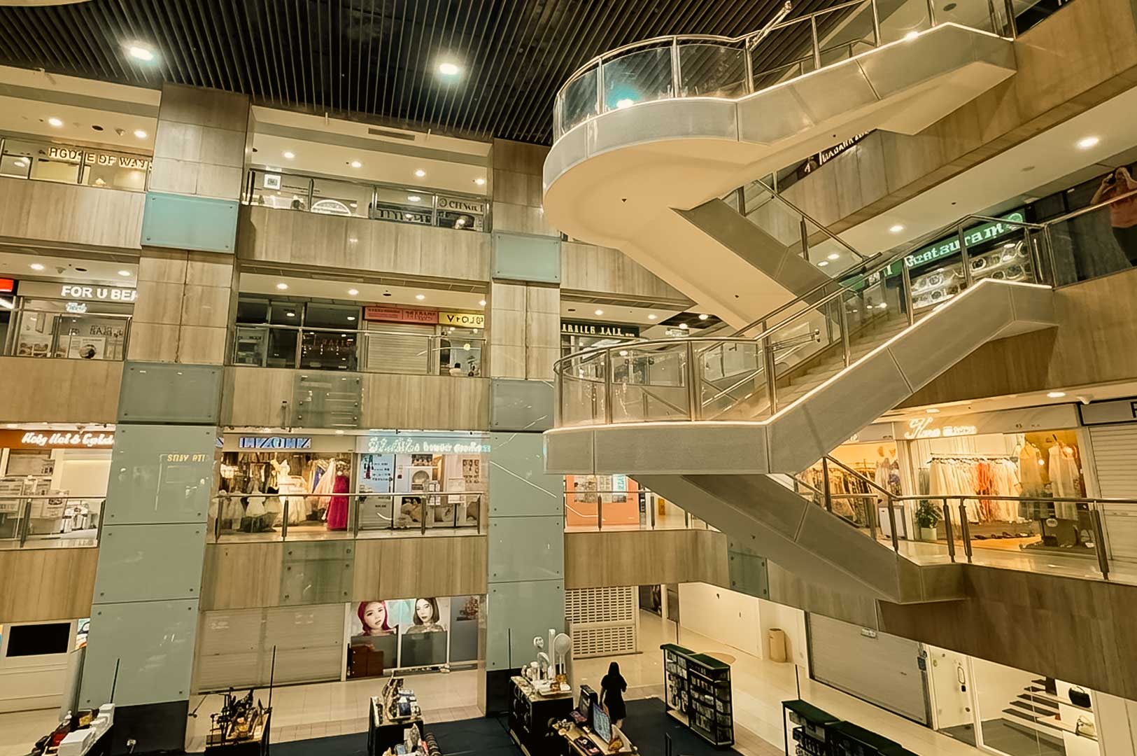 A low-angle view looking up at a multi-level indoor shopping mall atrium featuring sweeping helical staircases with glass railings, surrounded by tiers of boutique store fronts and kiosks.
