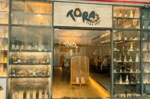 A wide exterior view of the Tora Tora Tora Japanese restaurant front, featuring glass doors and windows flanked by floor-to-ceiling display shelves filled with various sake bottles, with a wooden sign reading "TORA TORA TORA" above the entryway.