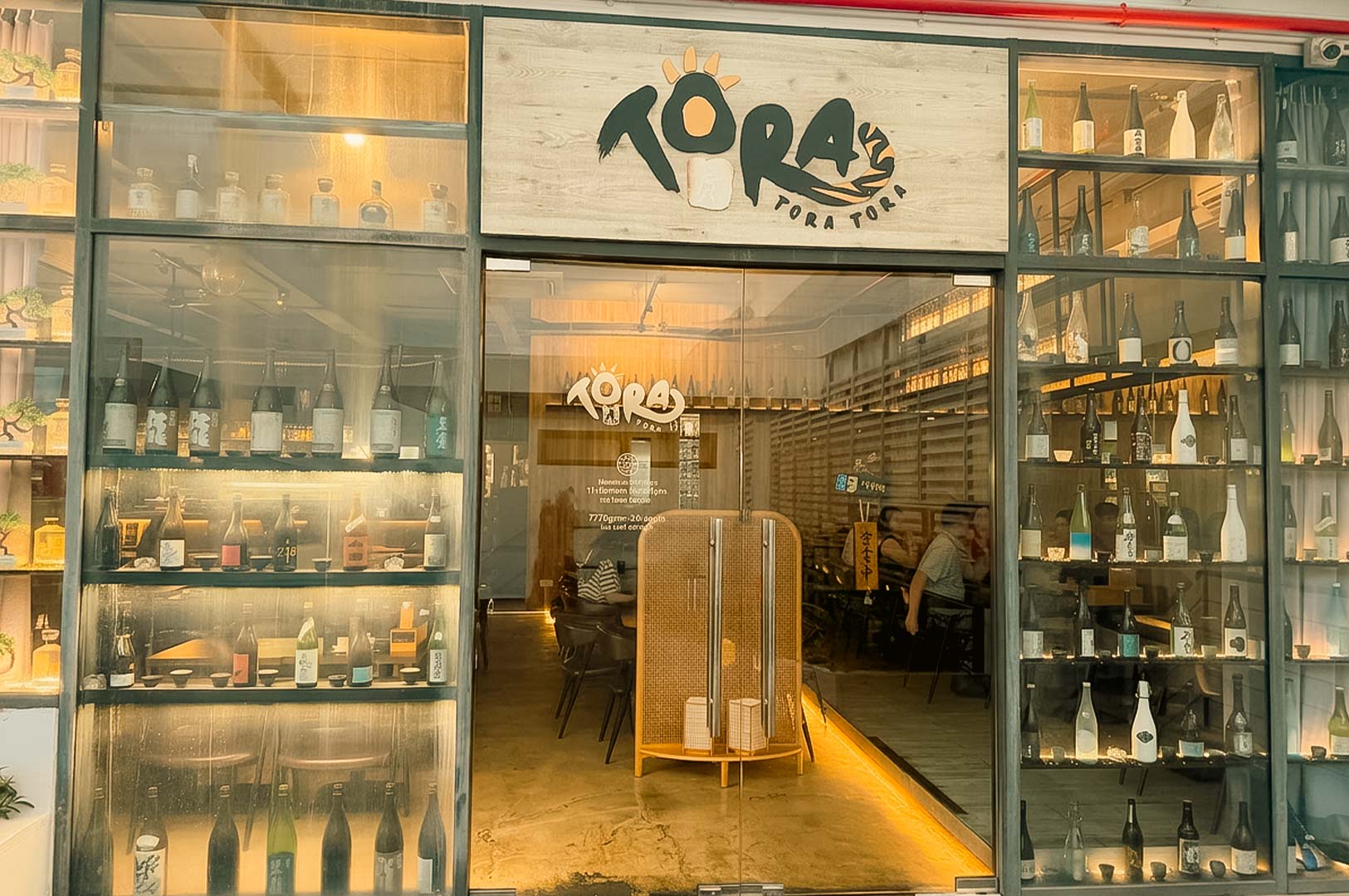 A wide exterior view of the Tora Tora Tora Japanese restaurant front, featuring glass doors and windows flanked by floor-to-ceiling display shelves filled with various sake bottles, with a wooden sign reading "TORA TORA TORA" above the entryway.