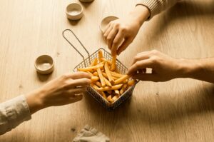 An overhead view of three people sharing french fries from a wire mesh serving basket on a light wooden table, surrounded by small dip ramekins.