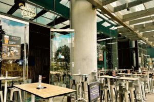 Modern open-air dining area outside Waa Cow!, with rows of wooden tables and metal chairs beneath a covered glass-and-steel canopy.