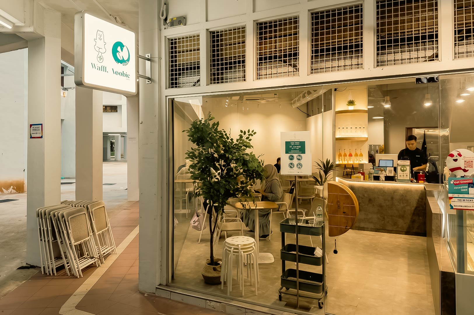 The image shows the exterior and interior storefront of a cafe named Waffl. Noobie, featuring a glowing sign with a bear illustration, glass walls looking into a brightly lit dining area with seating and a potted tree, and a counter space with a staff member visible inside. Stacked folding chairs are lined up on the walkway outside to the left.