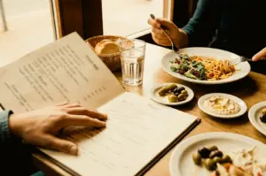Angle shot of two people dining at a wooden restaurant table next to a window. In the foreground, one person rests their hand on an open, printed menu. Across the table, another person uses a fork and knife to eat a plate of pasta with side salad. Small appetizers, including dishes of mixed olives, creamy dip, a bread basket, and a glass of water, are spread across the table.