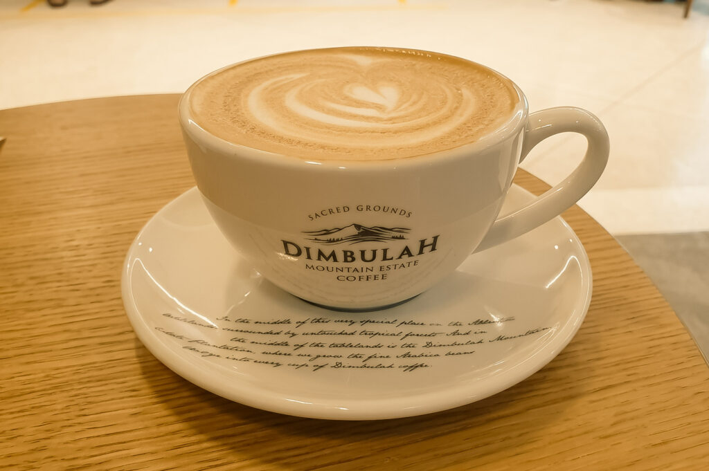 Close-up of a warm latte with delicate heart-shaped latte art, served in a white branded ceramic cup and saucer reading "Dimbulah Mountain Estate Coffee," resting on a light wooden tabletop.
