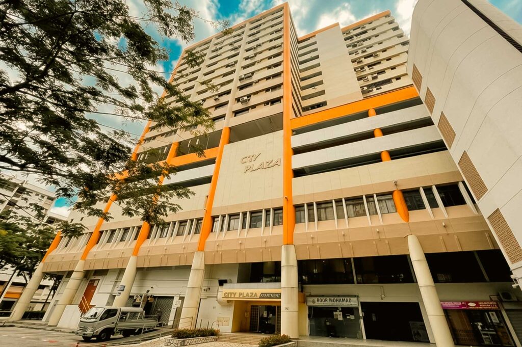 A low-angle exterior view of the City Plaza building with its distinctive orange and beige facade, featuring ground-floor storefront entrances and upper residential levels set under a bright blue sky.