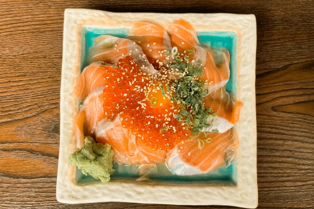 A top-down view of a salmon donburi bowl served in a square ceramic dish, featuring fresh salmon slices arranged in a ring, topped with bright orange ikura (salmon roe), a raw egg yolk, sesame seeds, chopped spring onions, and a dollop of wasabi on the side.