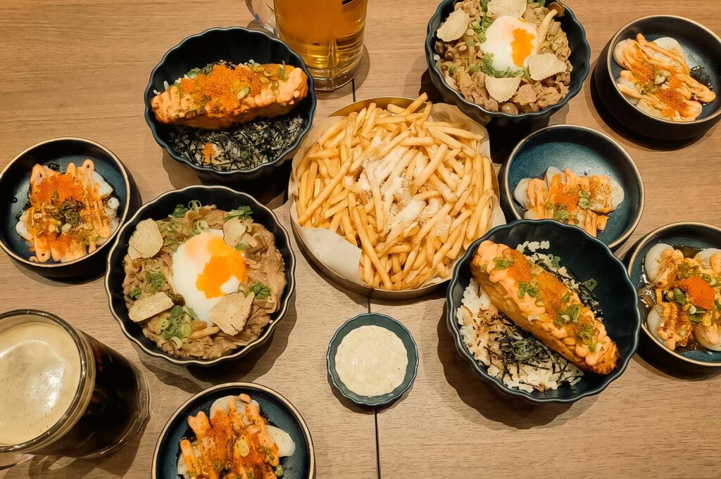 Overhead view of a Japanese-style spread featuring sushi bowls, beef bowls topped with soft eggs, crispy fries, assorted seafood dishes with spicy mayo and roe, and drinks on a wooden table.