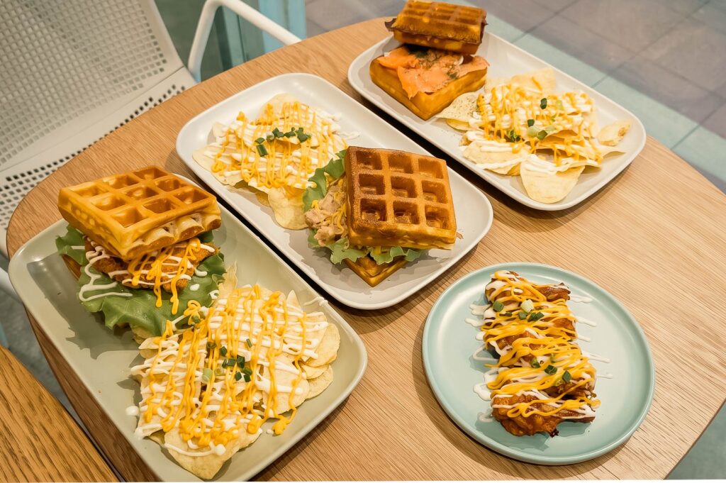 A wooden table at the cafe displays an assortment of savory waffle dishes, including golden-brown waffle burgers filled with ingredients like meat and lettuce, an open-faced waffle topped with smoked salmon, and a side plate of snacks, all served alongside crispy chips generously drizzled with cheese and white sauces.