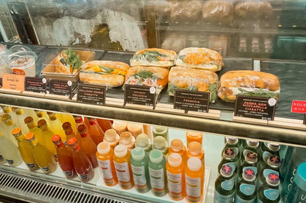 A display case at Dimbulah Coffee filled with fresh food options. The top shelf displays wrapped sandwiches, focaccia, ciabatta, and yogurt cups with price tags, while the bottom shelf is neatly lined with colorful bottled juices and San Pellegrino sparkling water.