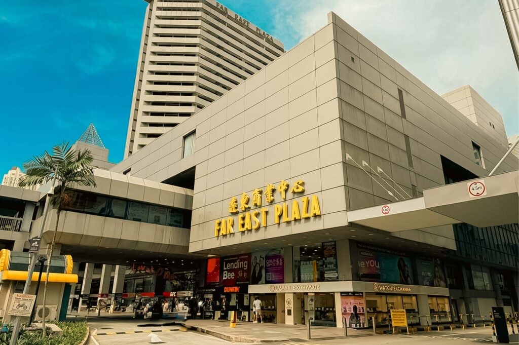 An exterior low-angle view of Far East Plaza featuring its prominent gold signage on a grey tiled facade, with ground-floor shops and a high-rise building towering in the background under a clear sky.