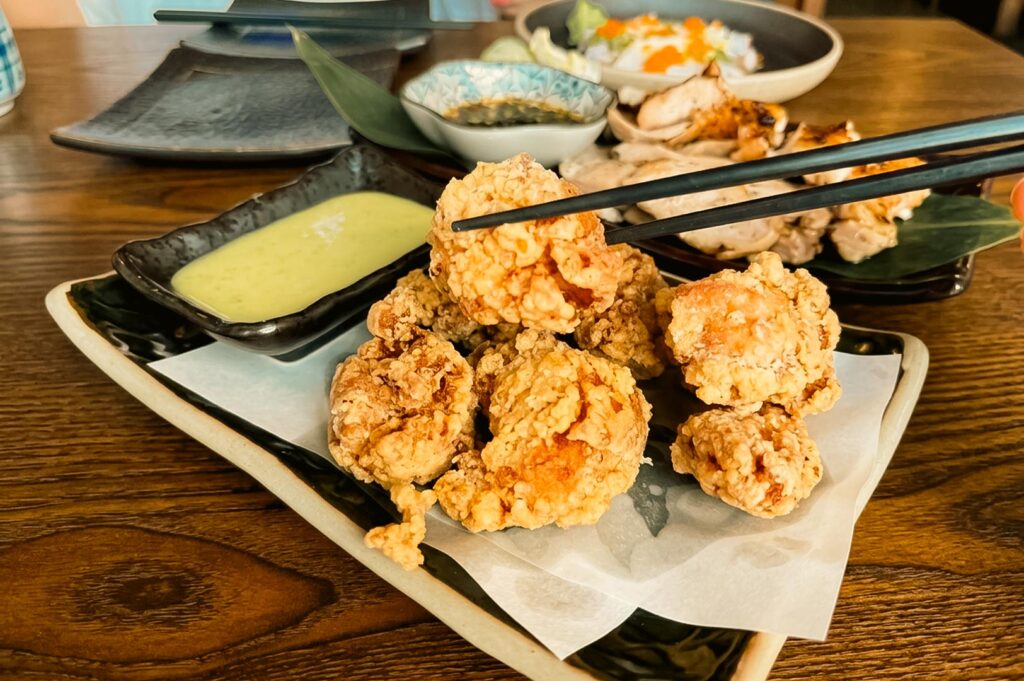 A pair of chopsticks holding up a piece of golden-brown Japanese chicken karaage from a plate, served with a side of creamy green dipping sauce and additional Japanese dishes in the background.
