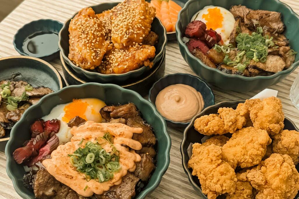 Close-up of a Japanese-style meal spread featuring beef rice bowls topped with soft eggs, green onions and spicy mayo, sesame-coated fried chicken, crispy chicken pieces, salmon slices, pickled vegetables, and dipping sauces.