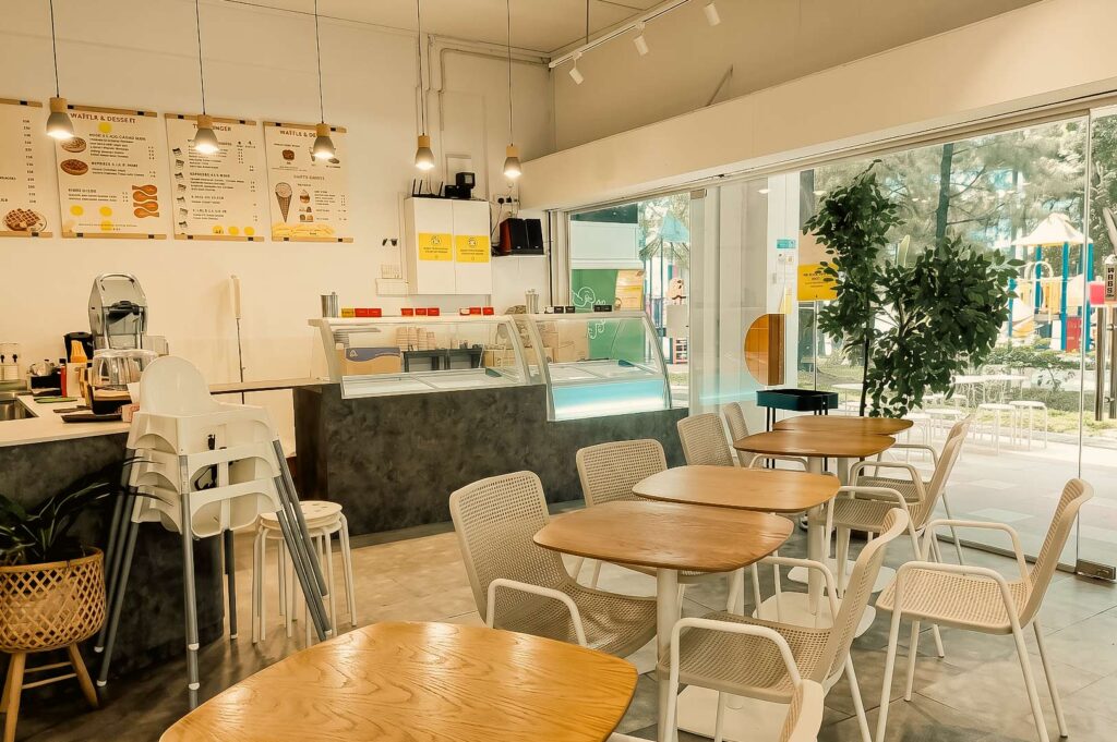 he image shows the interior of the cafe, featuring several wooden tables with white and beige chairs, a service counter with glass display cases, menu boards mounted on the wall, and large glass windows and doors that look out onto an outdoor playground area.