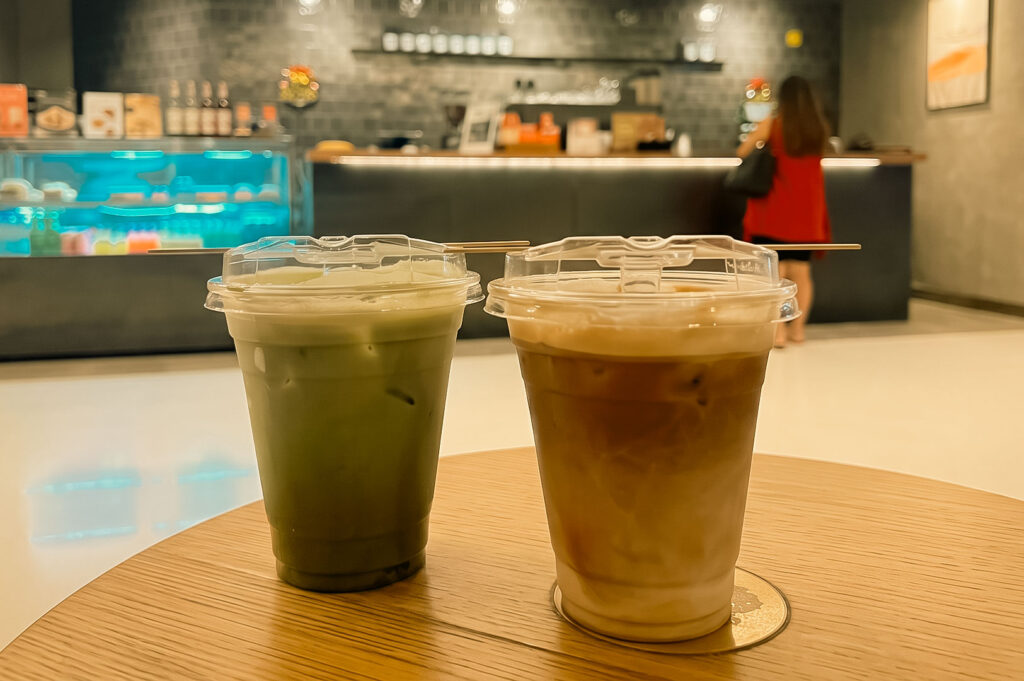 Alt Text Two iced beverages in clear plastic cups with sippy lids rest side by side on a light wood table, one featuring a green matcha latte and the other an iced coffee. In the blurred background, a cafe counter and a customer in a red top are visible.