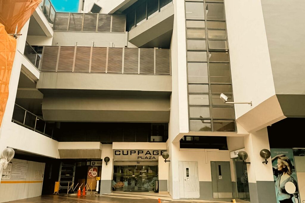 An exterior view of the Cuppage Plaza entrance showcasing modern architectural geometric white and grey concrete balconies, horizontal slatted screens, and glass panel windows.