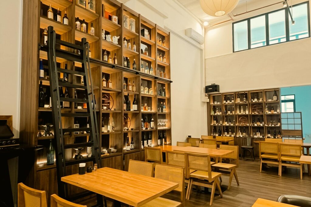 An interior view of Tora Tora Tora Japanese restaurant featuring high ceilings, wooden dining tables with woven rattan chairs, and a massive floor-to-ceiling wooden shelving unit filled with sake and liquor bottles, complete with a rolling black library ladder.