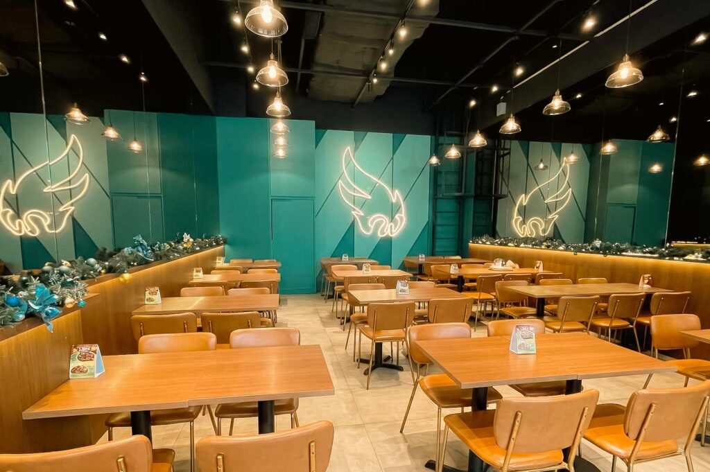 Spacious Japanese restaurant interior with rows of wooden tables and tan chairs, teal geometric walls, warm pendant lights, and illuminated Waa Cow! logos, decorated with festive ornaments.