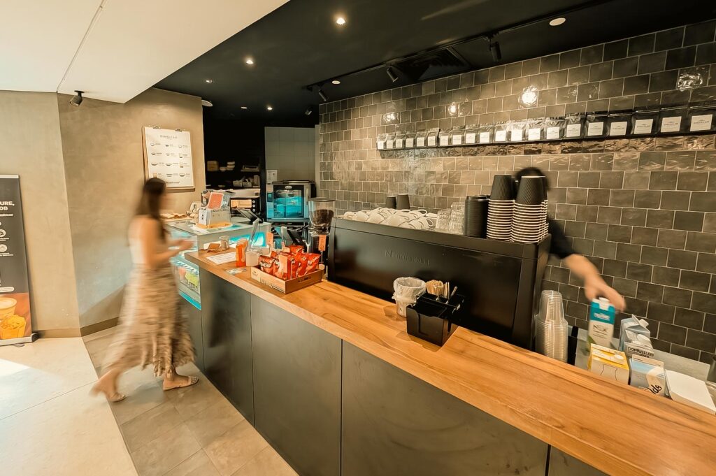 Wide shot of a modern cafe service counter featuring a warm wood top, sleek black lower paneling, and dark gray tiled accent walls. A espresso machine, stacked paper cups, and retail coffee bags are arrayed behind the counter while a customer walks past the order section.