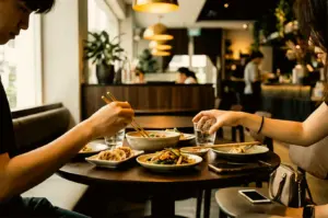 Two people sitting at a dark round table in a warmly lit cafe, sharing dumplings and noodle dishes during a meal.