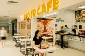 This wide-angle shot showcases the storefront entrance of Bao Er Cafe inside a brightly lit building hallway, dominated by large yellow lettering reading "BAO ER CAFE" above the main opening. The space features small white tables paired with round stools along the corridor where a customer dines, while staff work behind a long order counter on the right, backed by white tile walls, framed photos, and a menu board.