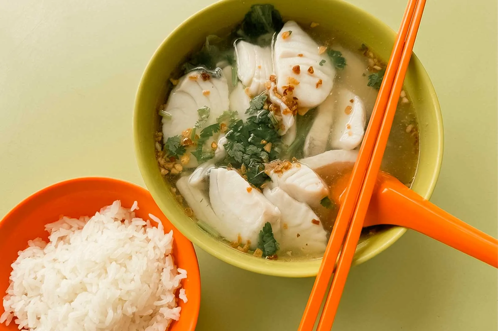 An overhead shot on a light green surface displays a bright yellow bowl filled with clear fish soup loaded with thick, tender slices of white fish, fresh cilantro, and golden fried garlic crisps. A pair of orange plastic chopsticks rests across the top of the bowl alongside a matching orange soup spoon, placed next to a small orange plastic bowl packed with steamed white rice.