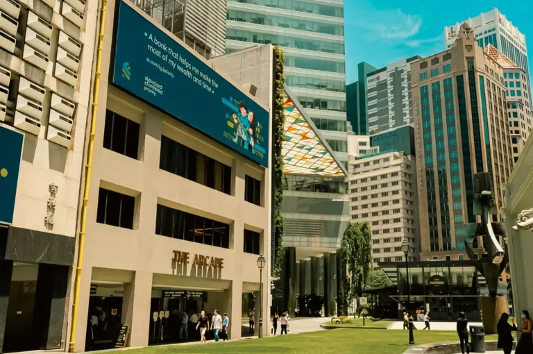 Street-level view of The Arcade entrance at Raffles Place surrounded by modern financial district skyscrapers and a green public plaza.