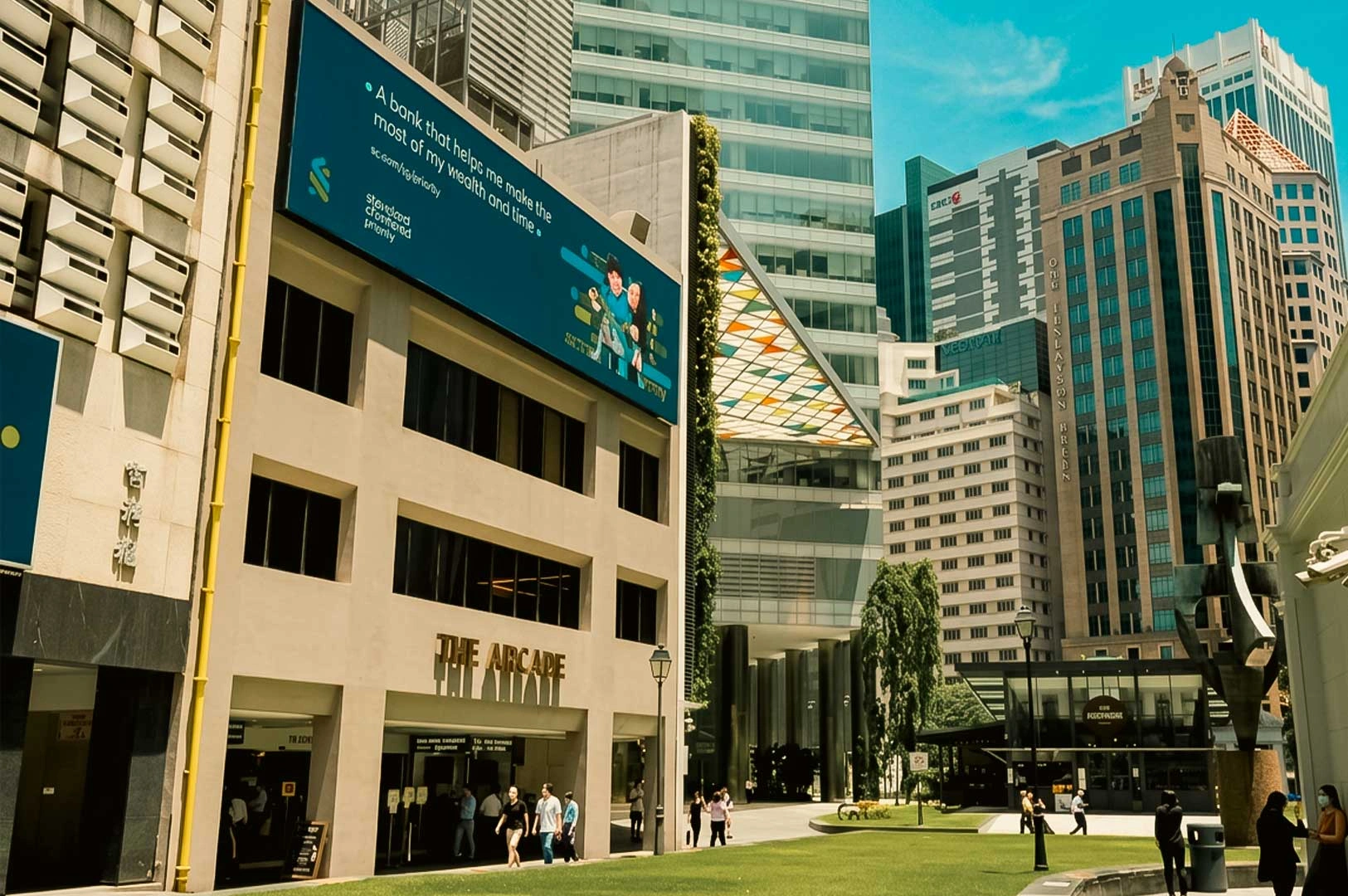Street-level view of The Arcade entrance at Raffles Place surrounded by modern financial district skyscrapers and a green public plaza.