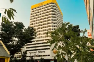 A low-angle shot showing the tall, white commercial building of Fortune Centre under a clear blue sky. The top section of the building features yellow trim with "FORTUNE CENTRE" signage, while the lower facade displays the building name in English and Chinese characters. Framing the foreground are vibrant green trees and lush foliage, giving a street-level perspective of the iconic building.