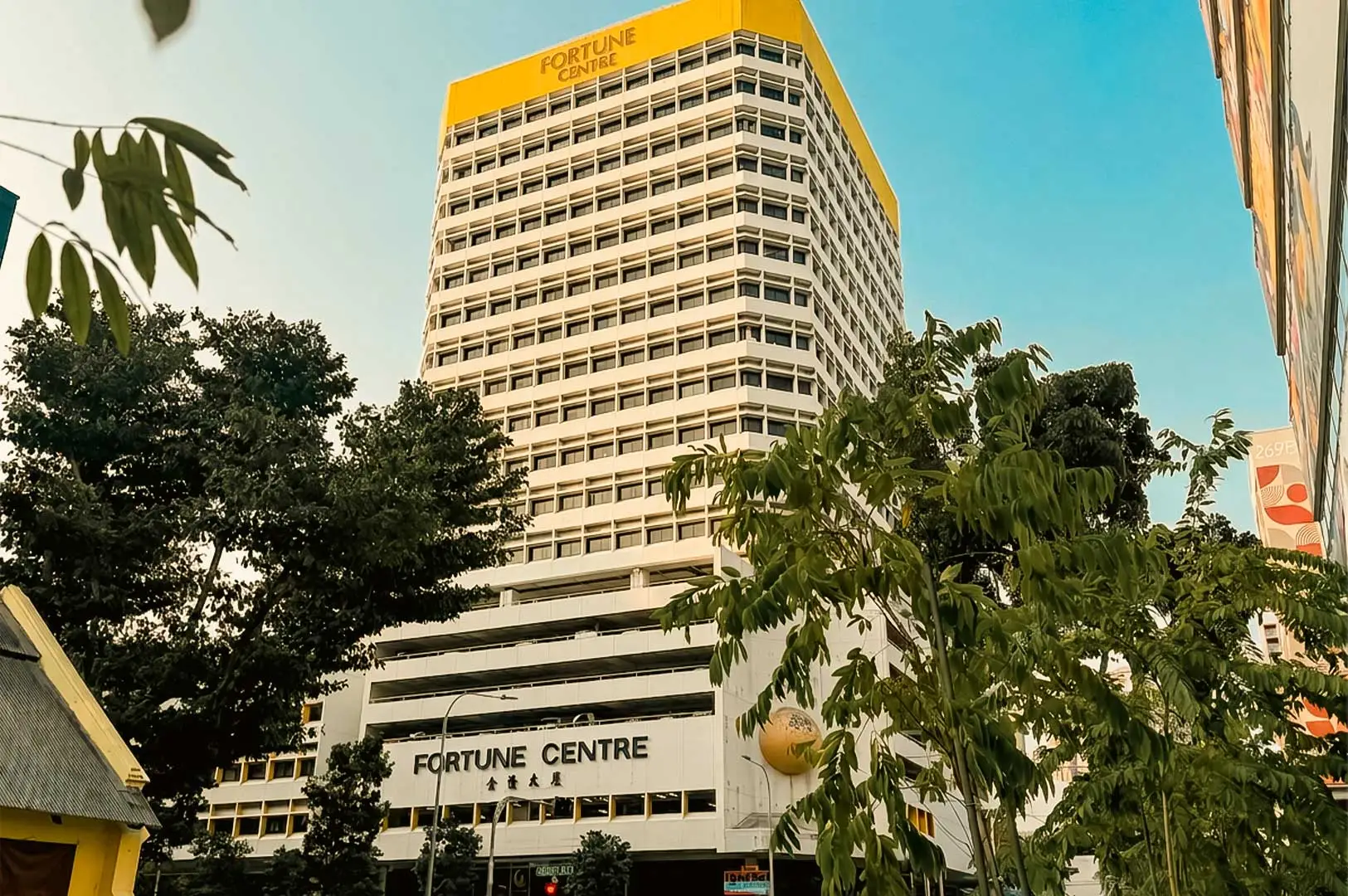 A low-angle shot showing the tall, white commercial building of Fortune Centre under a clear blue sky. The top section of the building features yellow trim with "FORTUNE CENTRE" signage, while the lower facade displays the building name in English and Chinese characters. Framing the foreground are vibrant green trees and lush foliage, giving a street-level perspective of the iconic building.