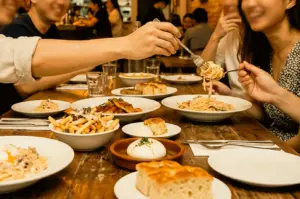 A hand reaching across a rustic wooden table filled with shared dishes—pasta, french fries, bread, and burrata—using a fork to take a bite of pasta straight from a friend's plate. A group of friends smiling and dining together in a warm, cozy restaurant setting.