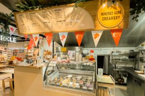 A bright ice cream shop counter featured under a large wooden sign displaying the name "HUNDRED ACRE CREAMERY" alongside a menu list, with decorative Singaporean flag pennants strung across the top. In the foreground, a glass display freezer showcases various pans of gelato, while small Winnie-the-Pooh plush toys sit on the counter next to paper cups and a ordering register.