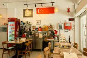 This interior shot captures the cozy dining space of Marie's Lapis Cafe, featuring light wooden tables set with woven condiment baskets and menus, accompanied by matching wooden chairs under warm hanging pendant lights. A red Coca-Cola beverage fridge stands on the left beside a service counter lined with shelves of knick-knacks and small decorations, while the back white wall displays framed artwork and a prominent Singapore flag hanging above the counter.