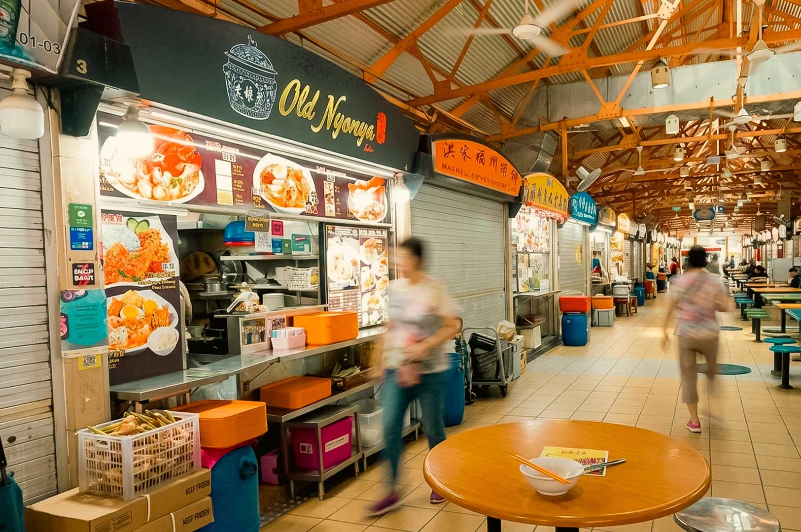 A wide shot of the Old Nyonya food stall located within the open-air layout of Maxwell Food Centre. The stall features a dark signboard with yellow lettering and Peranakan-style illustrations above display menus showcasing laksa and rendang dishes. A motion-blurred pedestrian walks past the stainless steel counter, while nearby dining tables, colorful stool seating, and high orange roof trusses line the background hawker aisle.