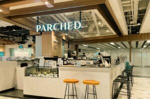 A modern, open-concept cafe kiosk named "PARCHED" features a illuminated teal sign mounted above a light-colored service counter equipped with a display case for bottled drinks and pastries. Wooden bar stools with black metal legs are arranged around the counter, set against a bright interior with warm wood ceiling accents and exposed overhead ductwork.