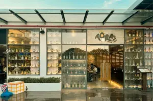 A modern restaurant storefront features floor-to-ceiling glass windows and doors beneath a sloped glass awning supported by dark metal frames. The signage above the entrance reads "TORA TORA TORA" in dark lettering next to a stylized sun logo. Large glass displays showcase neatly arranged bottles and small potted bonsai plants on illuminated shelves, while patrons are visible sitting at tables inside the warm-lit interior. Outside, a dark, polished patio floor holds a planter box with green foliage alongside a covered cooler and a single glass bottle resting against the base of the wall.