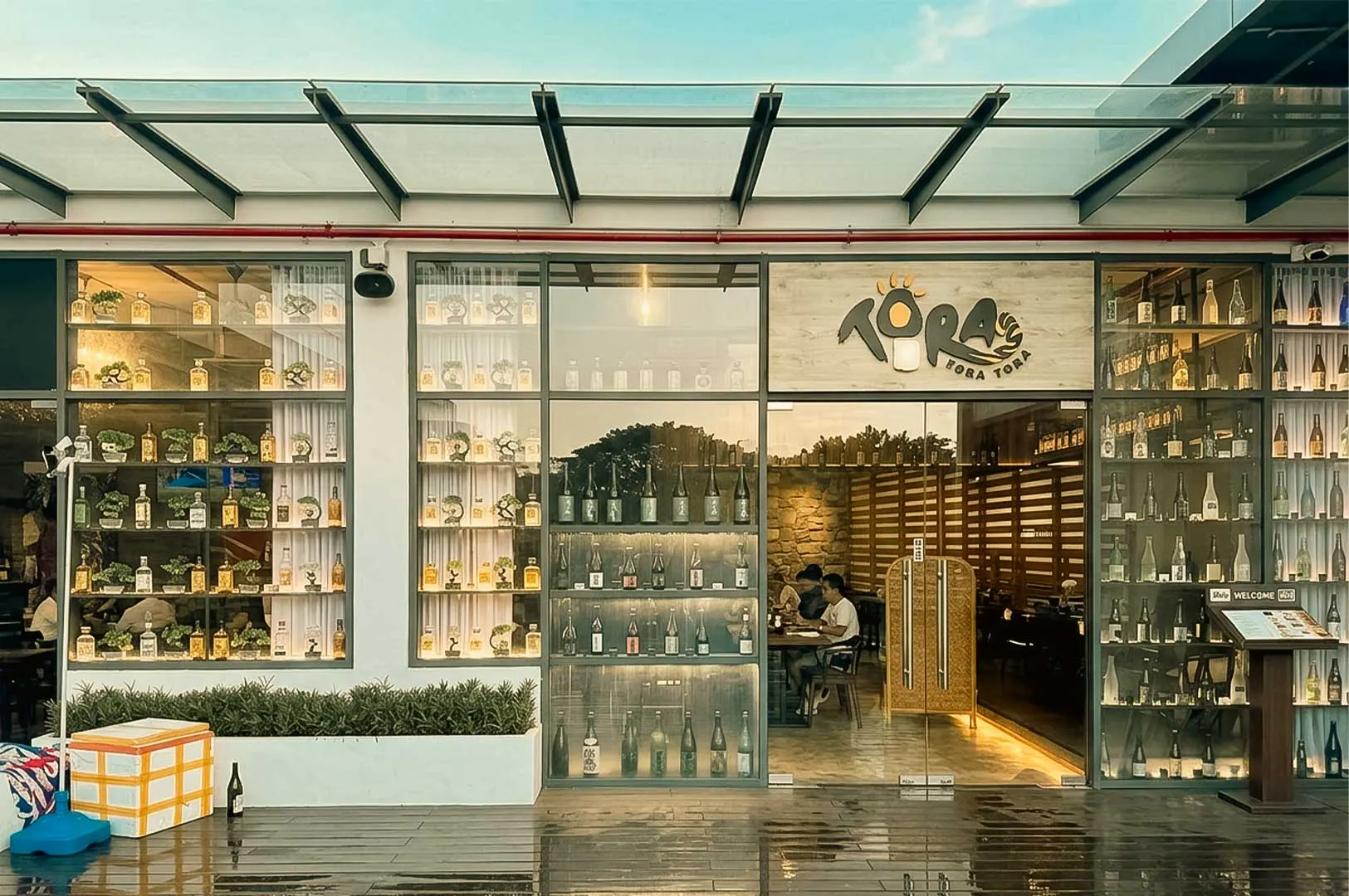 A modern restaurant storefront features floor-to-ceiling glass windows and doors beneath a sloped glass awning supported by dark metal frames. The signage above the entrance reads "TORA TORA TORA" in dark lettering next to a stylized sun logo. Large glass displays showcase neatly arranged bottles and small potted bonsai plants on illuminated shelves, while patrons are visible sitting at tables inside the warm-lit interior. Outside, a dark, polished patio floor holds a planter box with green foliage alongside a covered cooler and a single glass bottle resting against the base of the wall.