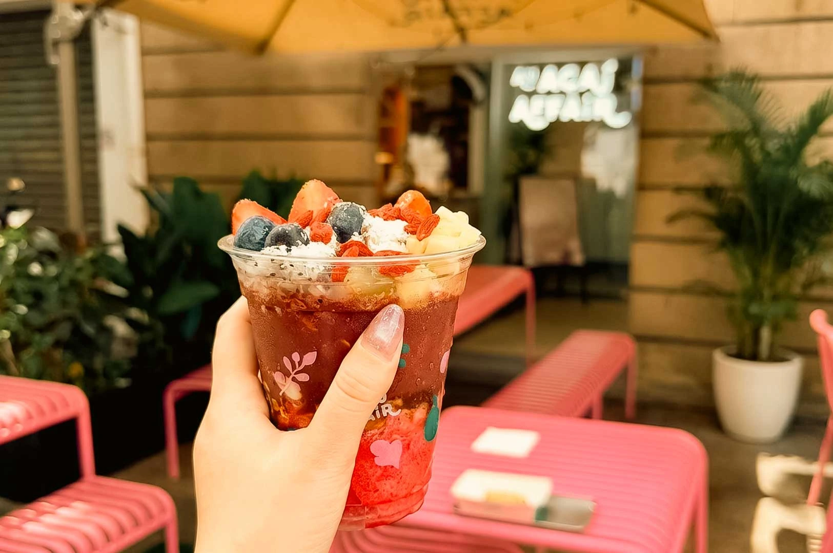 A close-up, first-person shot of a person holding a clear plastic cup filled with a layered açaí bowl from An Açaí Affair. The top of the cup is generously layered with fresh blueberries, sliced strawberries, banana chunks, goji berries, and a dusting of shredded coconut. The background features an outdoor cafe patio area with bright pink seating and potted green plants under a large umbrella canopy.
