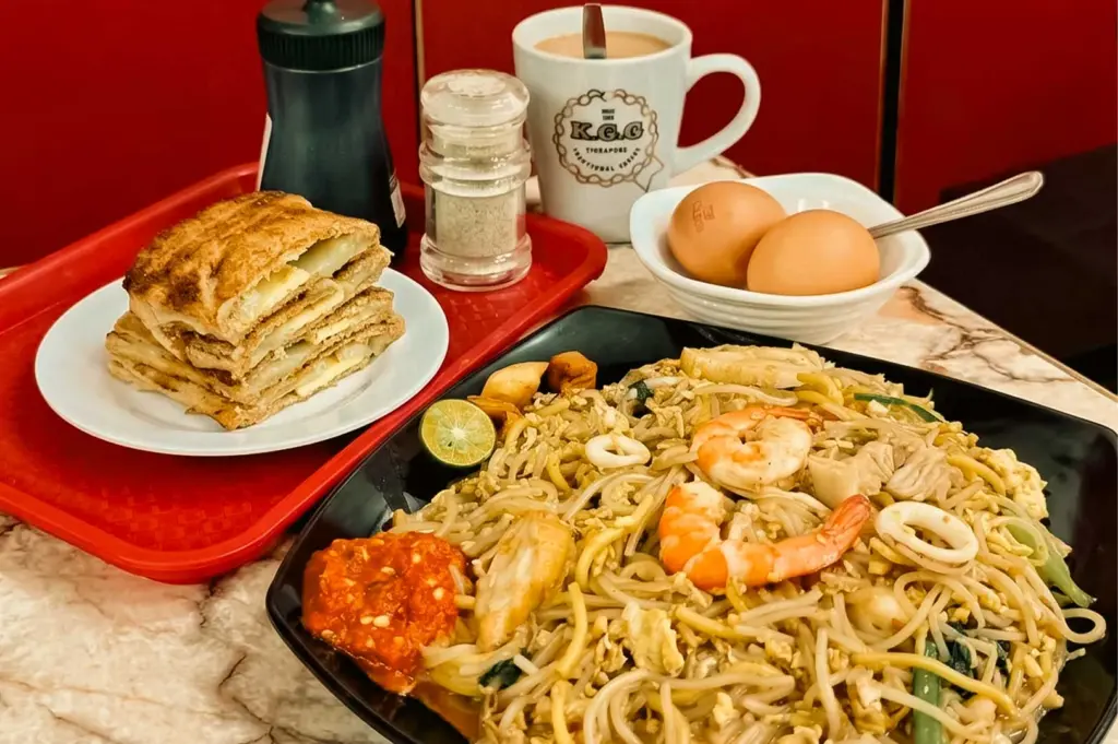 This close-up shot features a traditional Singaporean breakfast and lunch spread served at Bao Er Cafe, with a large black plate of Hokkien mee topped with prawns, squid rings, bean sprouts, and a side of red sambal chili in the foreground. In the background on a red plastic tray, stack of kaya toast slices sits on a white plate next to a dark soy sauce bottle, a pepper shaker, a white ceramic mug of hot milk tea, and a small white bowl holding two soft-boiled eggs.