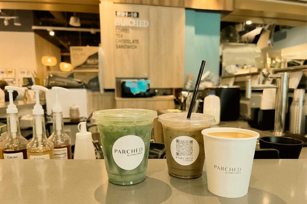 Three beverage cups branded with "PARCHED by PARCHMEN" logos sit lined up on a smooth cafe counter. The lineup includes an iced matcha latte in a clear plastic cup, an iced coffee beverage with a black straw, and a hot beverage in a white paper cup, with syrup dispensers and espresso station equipment visible in the softly blurred background.