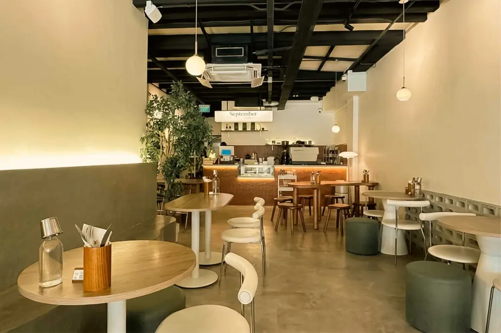 Minimalist cafe interior at September Coffee featuring light wooden tables, cream curved chairs, green ottomans, and a wooden coffee counter.